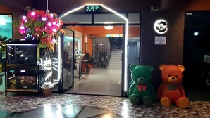 A hotel entrance with a digital clock displaying 19:28:04, two teddy bears, and a decorative plant with lights. - Lert Sri Hotel (Suphan Buri)