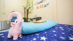 A room with a bed covered in a blue blanket with white stars, a pink stuffed animal, a blue pillow, a wall with a tree and text, and a small table with a chair. - Jumbo Villa Homestay and Resort (Wiset Chai Chan)