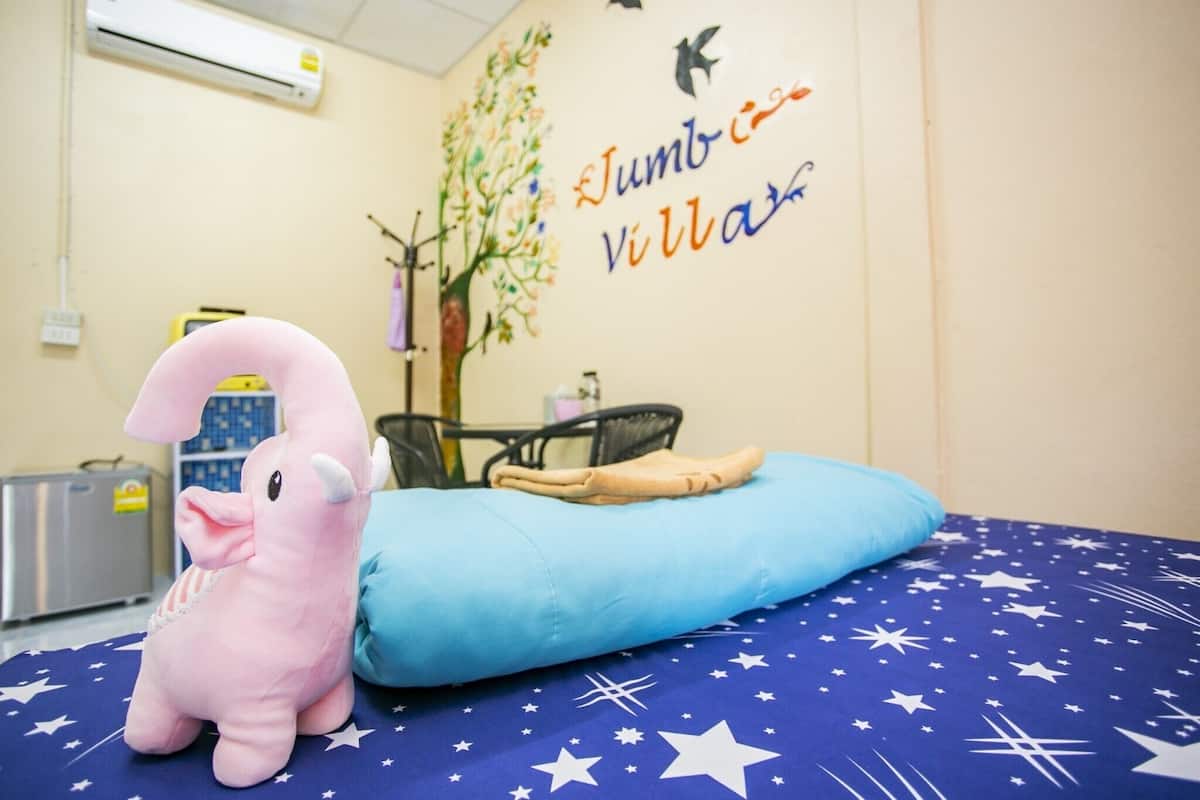 A room with a bed covered in a blue blanket with white stars, a pink stuffed animal, a blue pillow, a wall with a tree and text, and a small table with a chair.