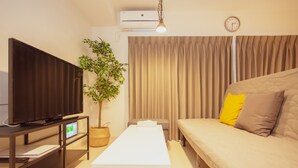 Et moderne hotelværelse med et fladskærms-tv, en plante, en sofa med puder, et hvidt bord og en gardin.