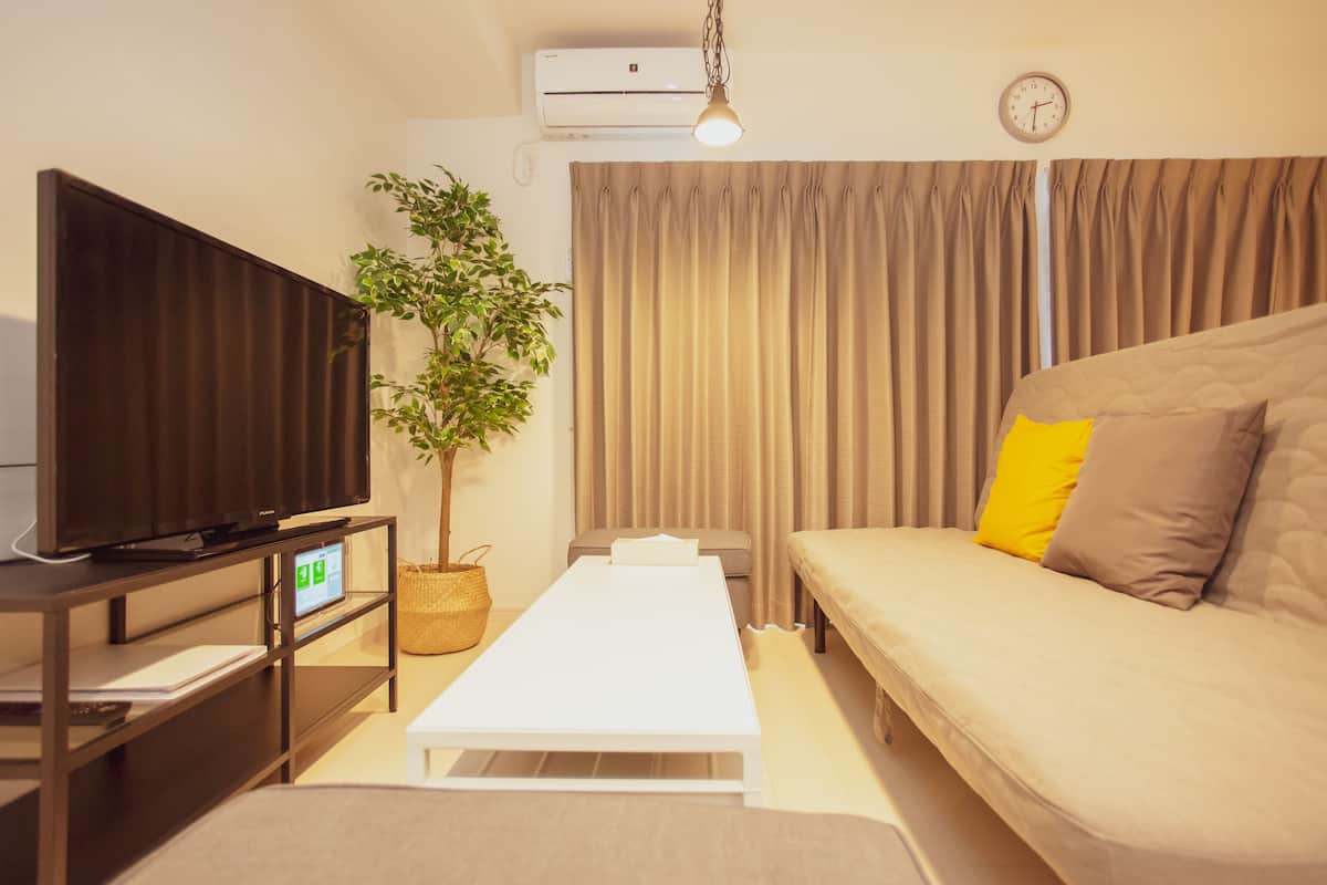 A modern hotel room with a flat-screen TV, a plant, a sofa with cushions, a white table, and a curtain.