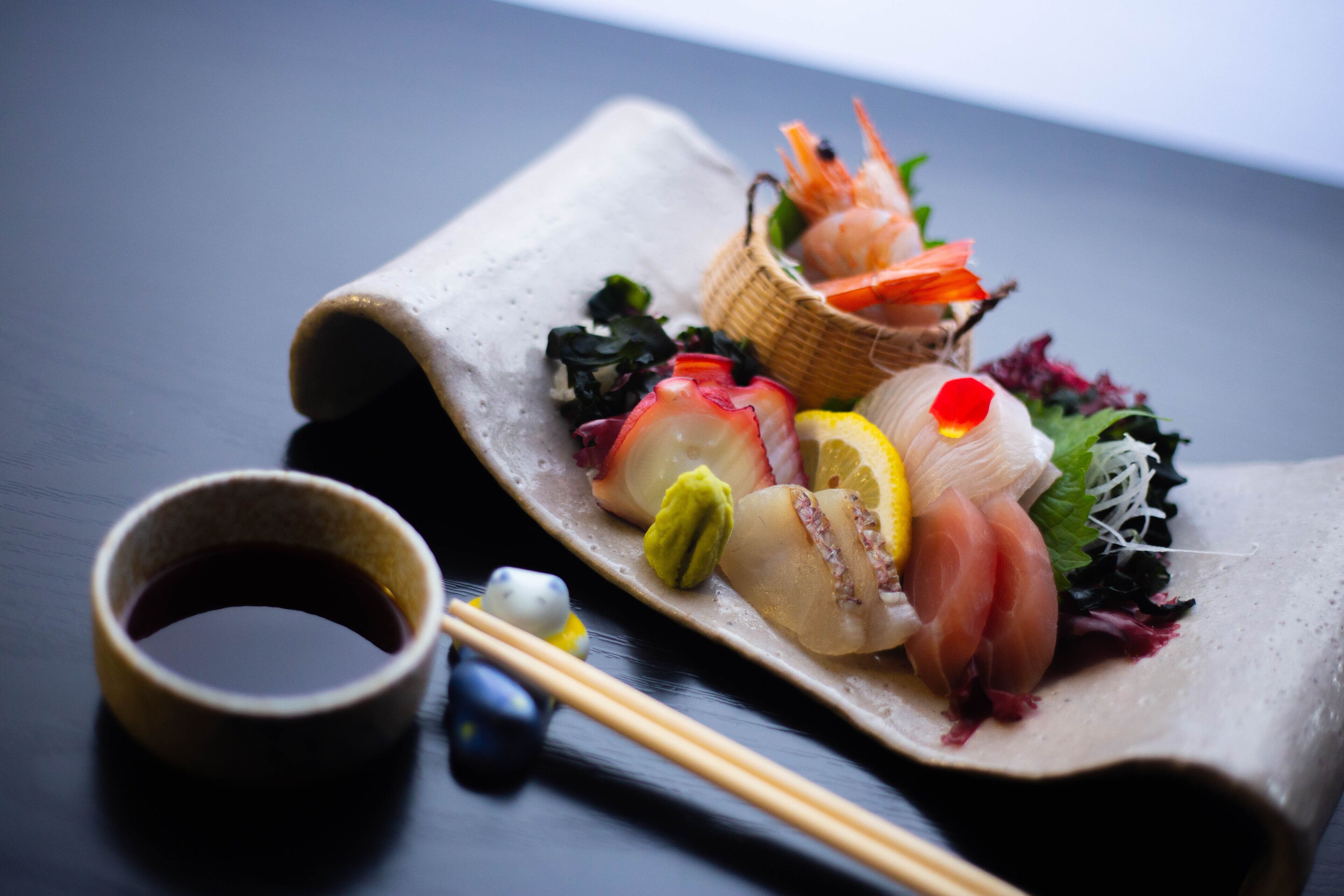 A sushi platter with various types of fish, vegetables, and a small basket of shrimp on a dark table.