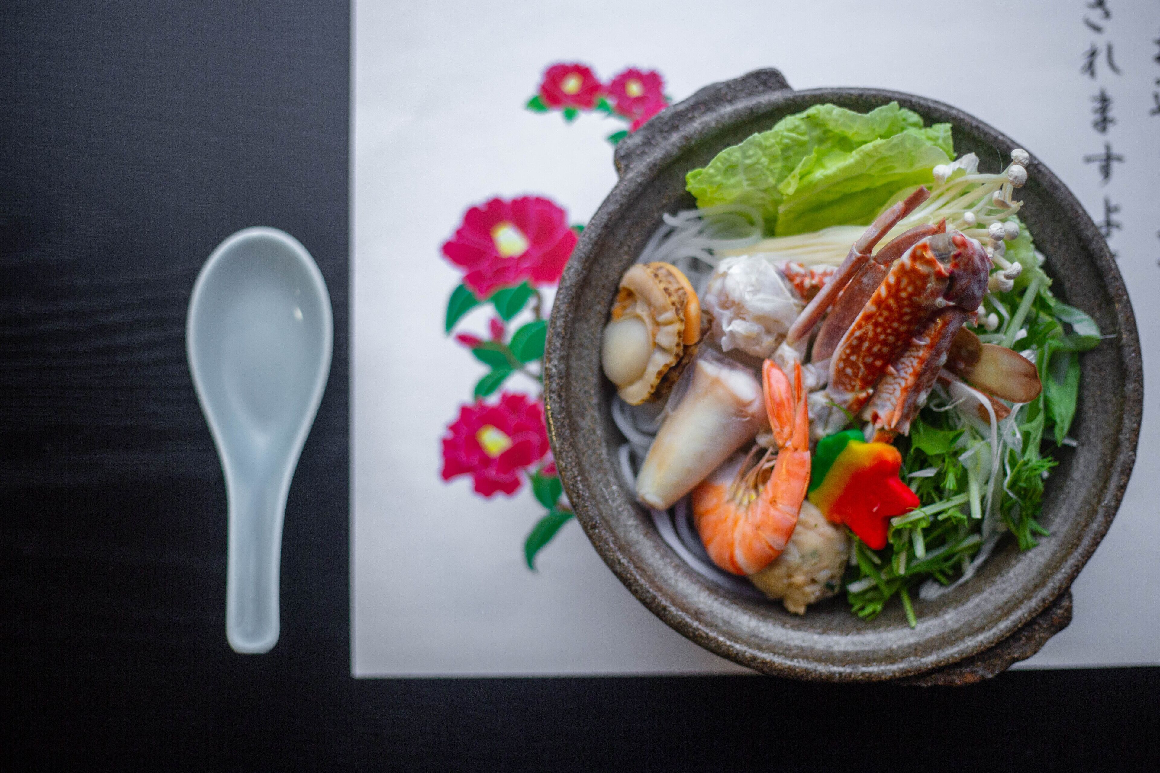 A bowl of seafood with vegetables and a white spoon on a black surface with floral decorations.