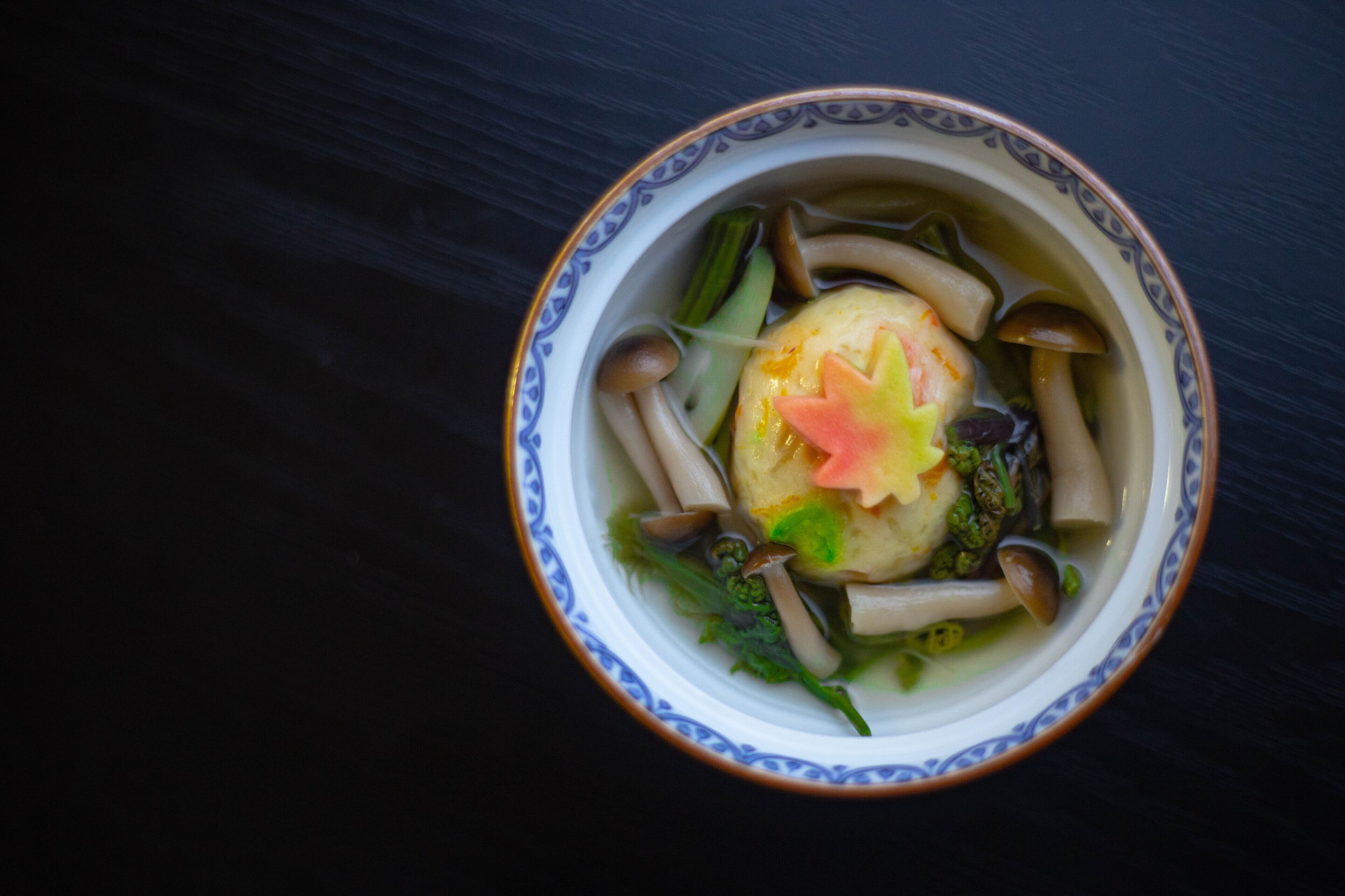 A bowl of soup with various vegetables and a dumpling, served in a decorative bowl on a dark surface.