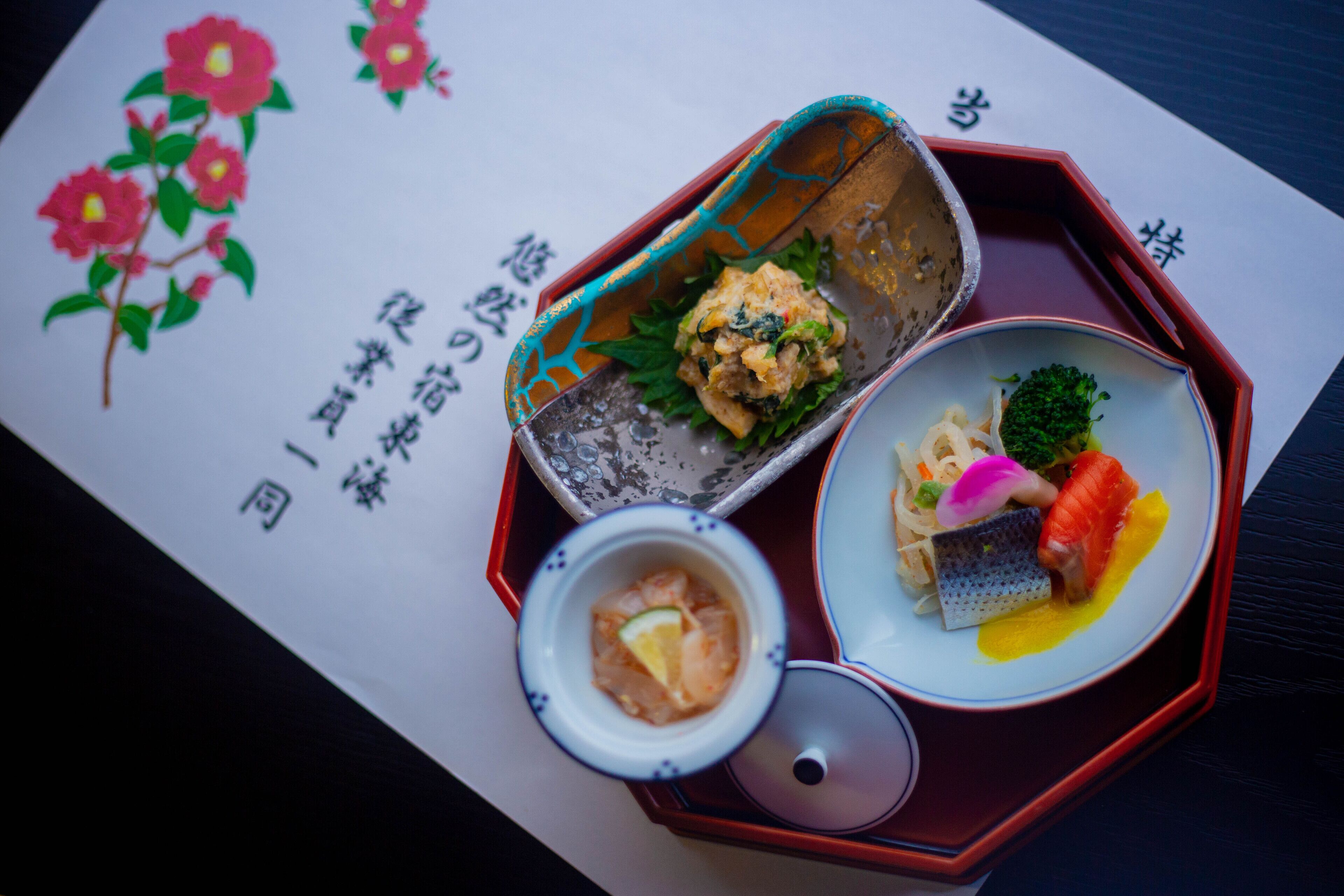 Comida tradicional japonesa servida en una bandeja de madera con diseño floral, que incluye un tazón de sopa, un plato con pescado y verduras, y una fuente con fideos y pescado.