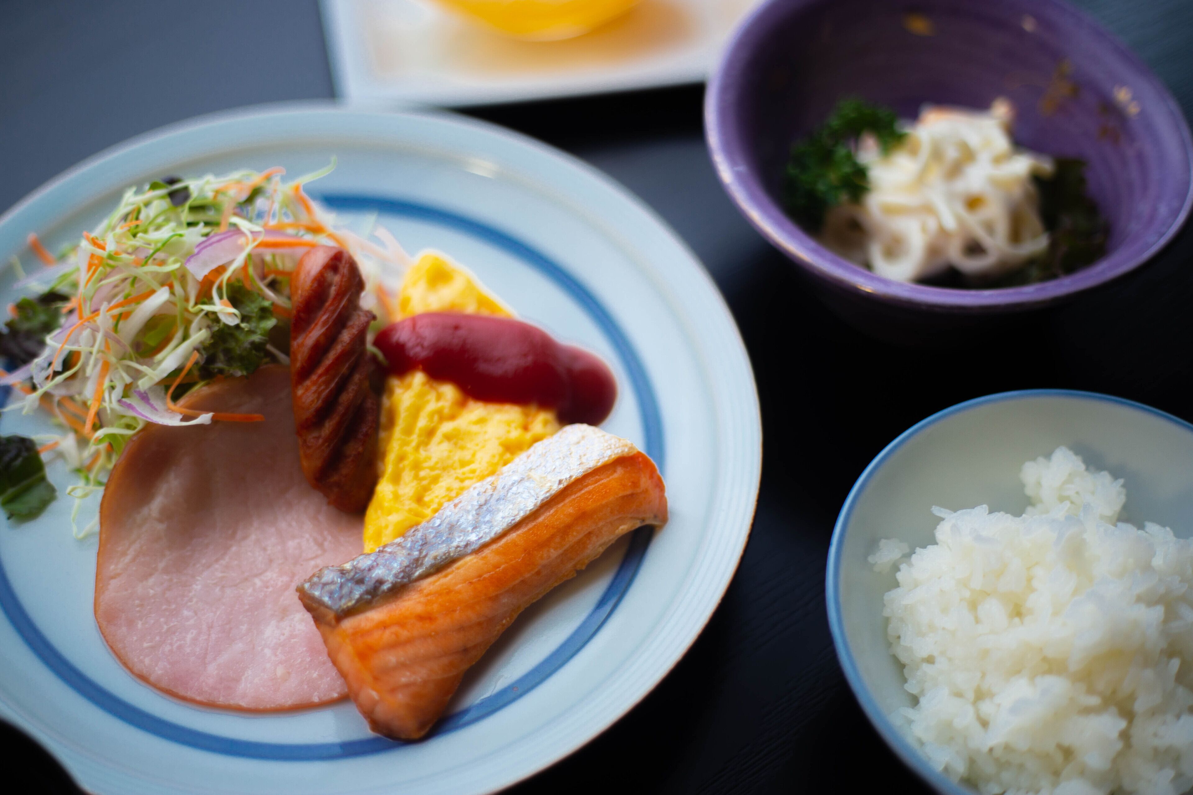 Un plato con salmón, jamón y una ensalada, un tazón de arroz y un tazón de fideos.