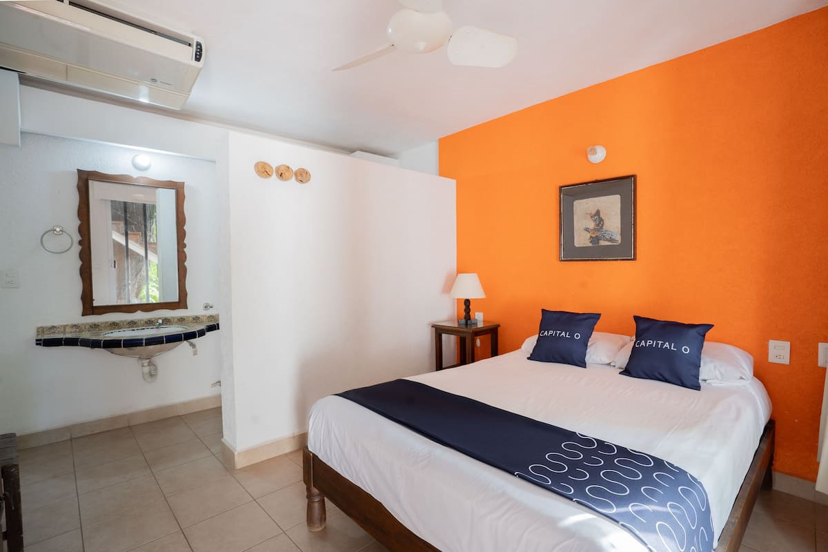 A hotel room with a bed, a nightstand with a lamp, an air conditioning unit, a mirror, and a sink.