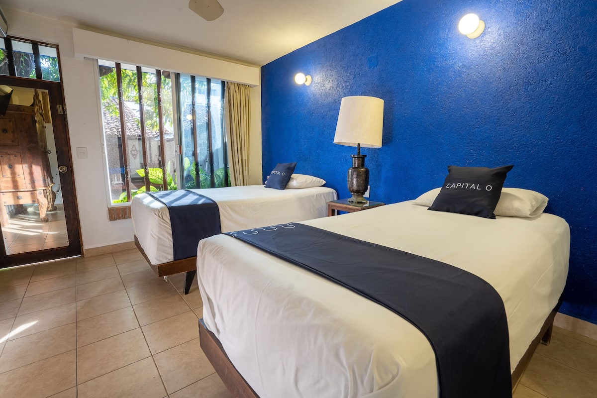 A hotel room with two beds, a blue accent wall, a window with curtains, and a lamp on a bedside table.