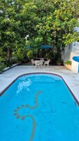 Una piscina con borde de azulejos, rodeada de exuberante vegetación y una mesa blanca para exteriores con sillas.