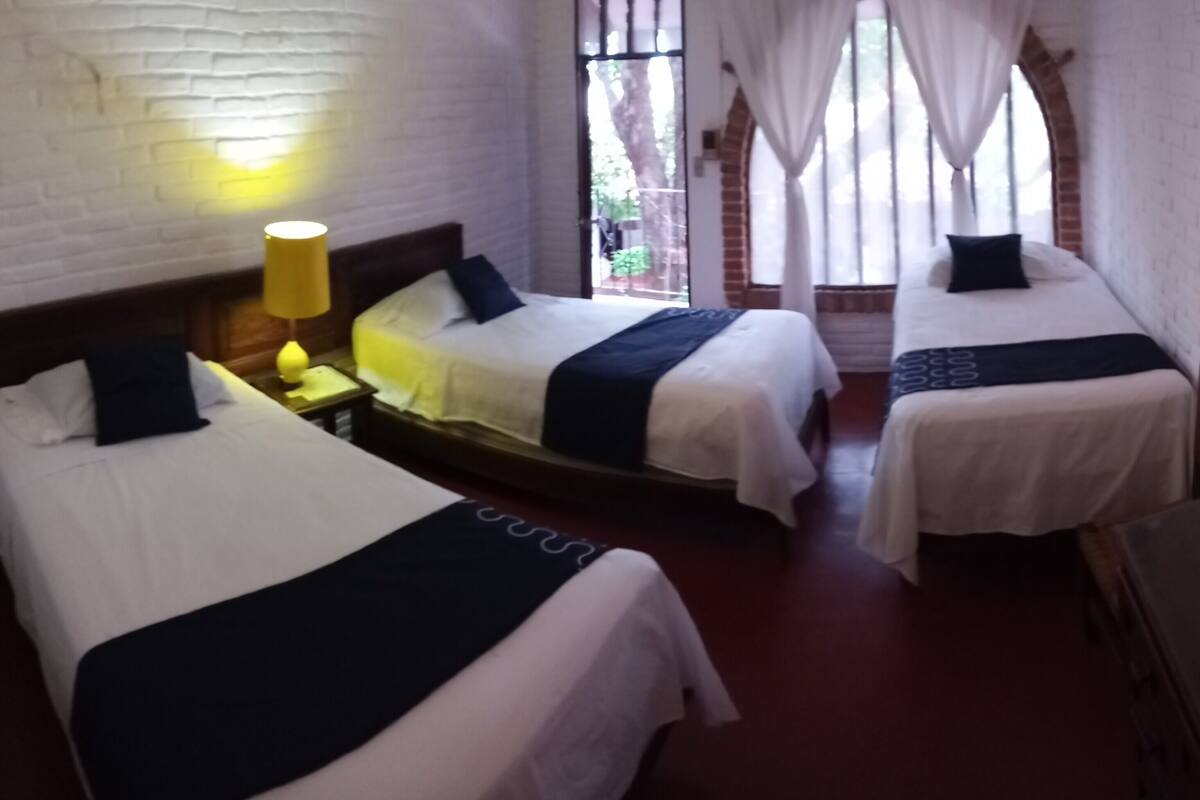 A hotel room with three single beds, a wooden nightstand with a lamp, and a brick wall.