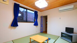 A room with a wooden ceiling, a window with blue curtains, a wooden table, a television on a cabinet, and a tatami mat floor. - Pension Sea Forest (Yakushima)