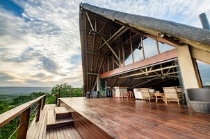 Una terraza de madera con vistas al paisaje, un amplio espacio abierto con asientos y una estructura de techo de paja.