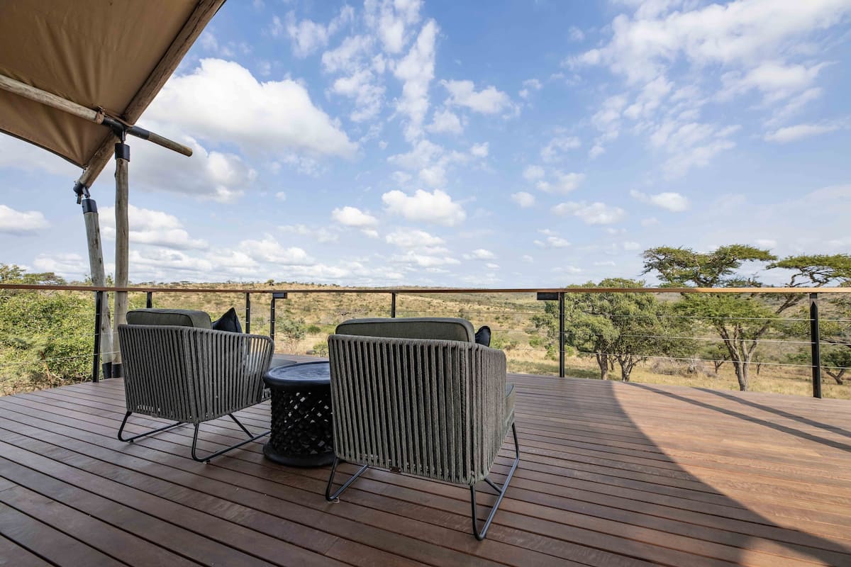 A wooden deck with two wicker chairs and a small round table, overlooking a natural landscape with sparse trees and a clear sky.