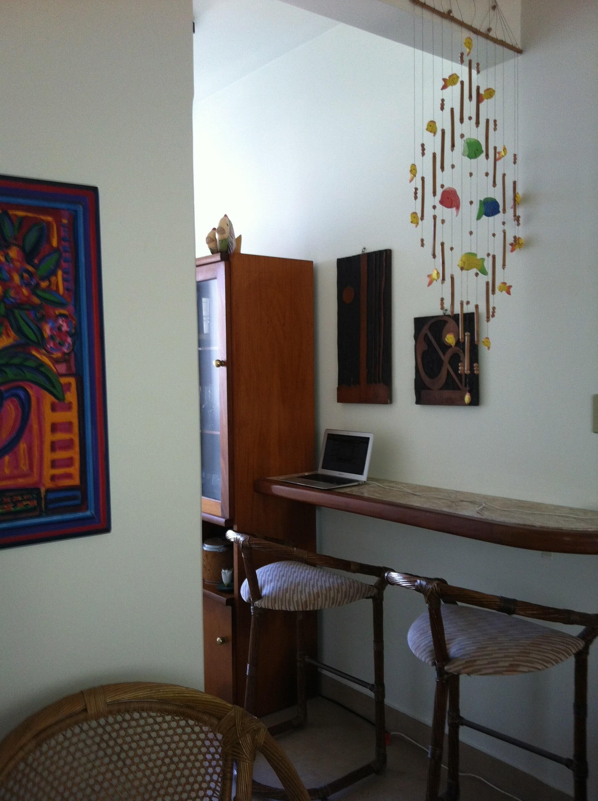A room with a desk, laptop, two wicker chairs, a decorative hanging, and a colorful painting.