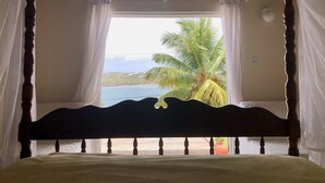 Un lit avec une tête de lit, des rideaux et une vue sur un palmier et l’eau.