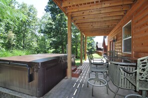 Ein überdachter Patio-Bereich mit Whirlpool, Sitzgelegenheiten im Freien und Blick auf ein bewaldetes Gebiet.