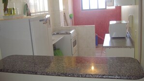 A compact kitchen with a granite countertop, a refrigerator, a microwave, and a window with blinds. - Guarujá-Enseada - 1 Bedroom Apartment with Pool (Guarujá)