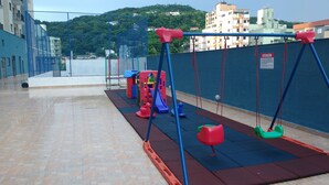 A children's play area with swings and a slide, surrounded by a blue fence and a red safety mat. - Guarujá-Enseada - 1 Bedroom Apartment with Pool (Guarujá)