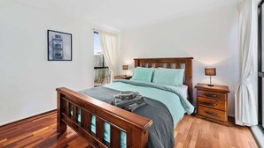 Un dormitorio con cama de madera, mesita de noche, cómoda, cuadro en la pared y ventana con cortinas.