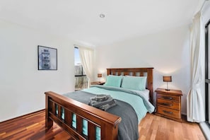 Une chambre à coucher avec un lit en bois, une table de chevet, une commode, un tableau encadré au mur et une fenêtre avec des rideaux.
