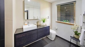 A modern bathroom with a dark vanity, a white sink, a mirror, and a toilet. There is a window with blinds and a small plant on the vanity.