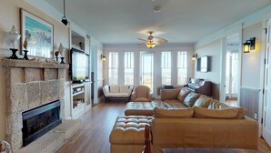 Ein geräumiges Wohnzimmer mit Kamin, einer großen Couch, einem Klavier und Blick auf den Strand durch die Fenster.