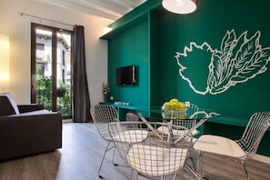 A modern room with a green accent wall, a dining area with wireframe chairs, a sofa, and a TV mounted on the wall.