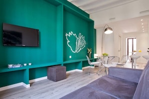 A modern living room with a green accent wall, a flat-screen TV, a dining area with a round table and chairs, and a grey sofa.