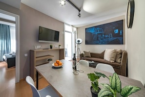 En moderne stue med et fladskærms-tv, en sofa, et spisebord med en skål frugt og et vinglas, samt en potteplante.