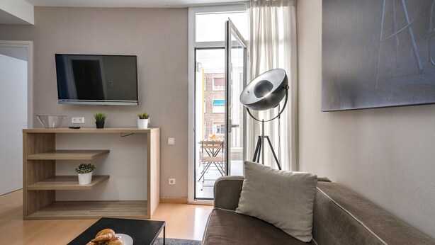 A modern living room with a flat-screen TV, a sofa, a coffee table with croissants, and a balcony with a view of buildings. - Cool Jazz 44 - Habitat Apartments (Barcelona)