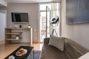 Un salon moderne doté d’un téléviseur à écran plat, d’un canapé, d’une table basse avec des croissants, et d’un balcon offrant une vue sur les immeubles.