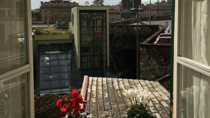 Une vue depuis une fenêtre donnant sur un paysage urbain, avec un mélange de bâtiments historiques et modernes, un mur de pierre et une zone pavée où sont garées des voitures.
