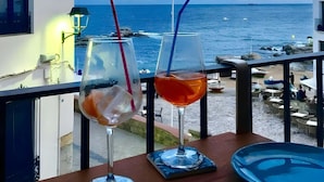 Uma varanda com duas bebidas, vista para o mar e uma pequena mesa com um prato azul.