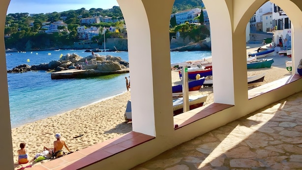 Vista da praia por uma galeria coberta com arcos, praia arenosa, barcos e edifícios ao fundo.