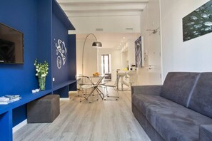 A modern living room with a grey sofa, a dining area with a table and chairs, a TV mounted on the wall, and a blue accent wall.