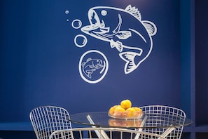A dining area with a table and chairs, a wall with a fish mural, and a bowl of fruit on the table.