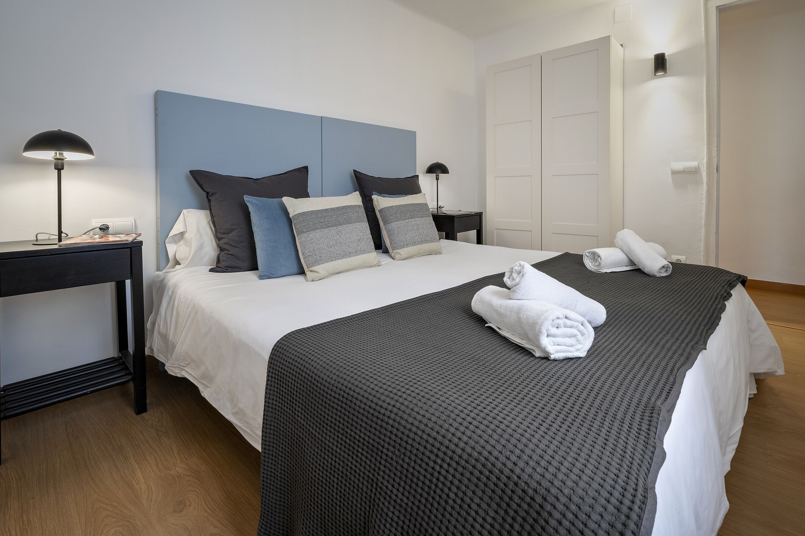 Ein ordentlich gemachtes Bett mit einer grau-weißen Tagesdecke, zwei grauen und schwarzen Kissen und zwei gefalteten weißen Handtüchern.