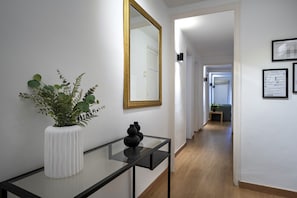 A hallway with a white wall, a mirror with a gold frame, a white vase with green plants, and a black vase on a glass table.