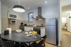 A modern kitchen with a round dining table, chairs, a bowl of fruit, cups, and a glass of orange juice.