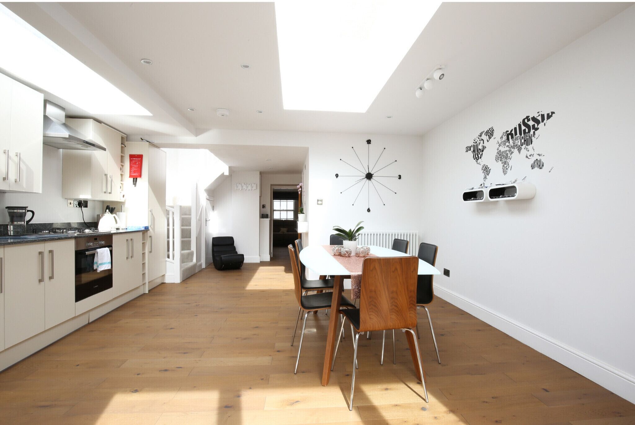 A modern kitchen with white cabinets, a dining area with a wooden table and chairs, and a world map mural on the wall.