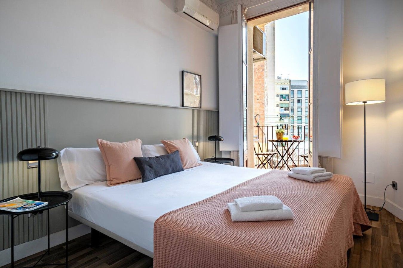 A hotel room with a bed, bedside tables, a balcony with a table and chairs, and a view of buildings.