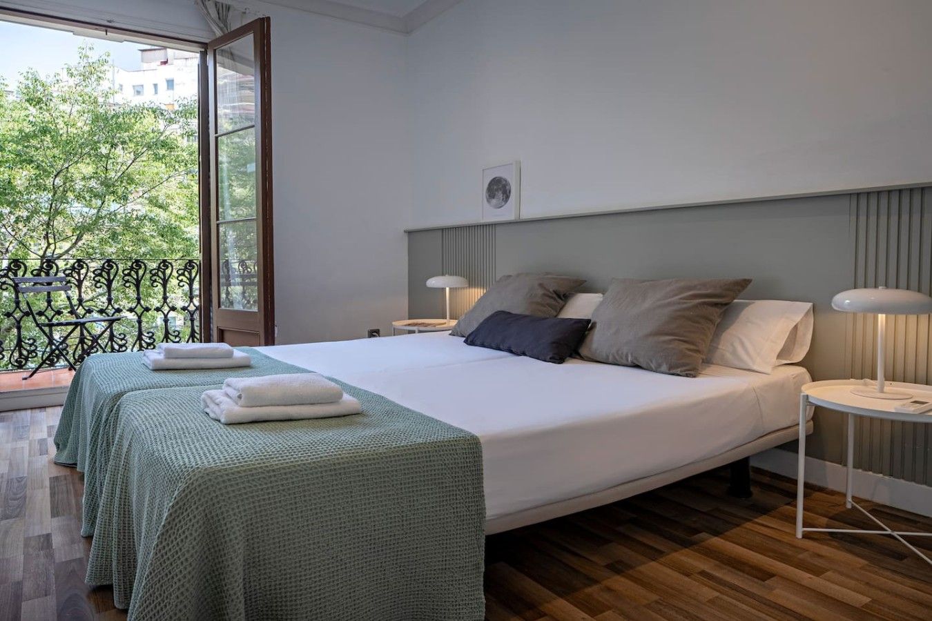 Ein Hotelzimmer mit einem großen Bett, zwei Nachttischen, einem Balkon mit schmiedeeisernem Geländer und Blick auf Bäume und Gebäude.