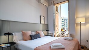A hotel room with a bed, bedside tables, a lamp, a window with a view of buildings, and a balcony with a table and chairs. - Barcelona Balconies 12 - Habitat Apartments (Barcelona)