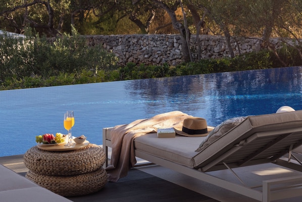 A poolside lounge with a reclining chair, a woven stool, a book, a hat, and a glass of orange juice.
