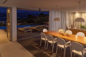 A modern dining area with a wooden table and white chairs, a view of a pool and mountains, and a glass door leading to an outdoor seating area.