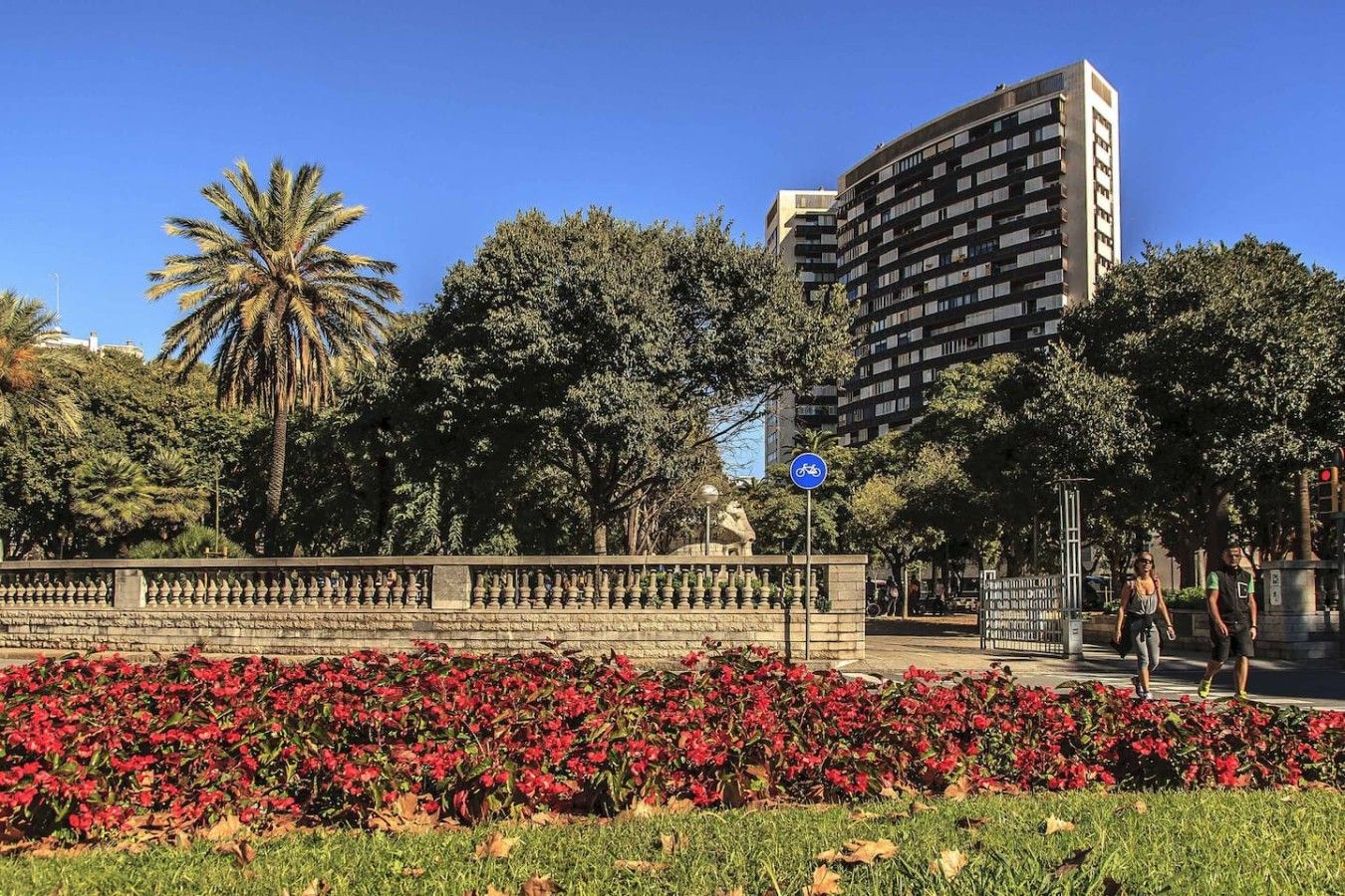 Un parc amb un mur de pedra, flors vermelles i un senyal blau de bicicleta.