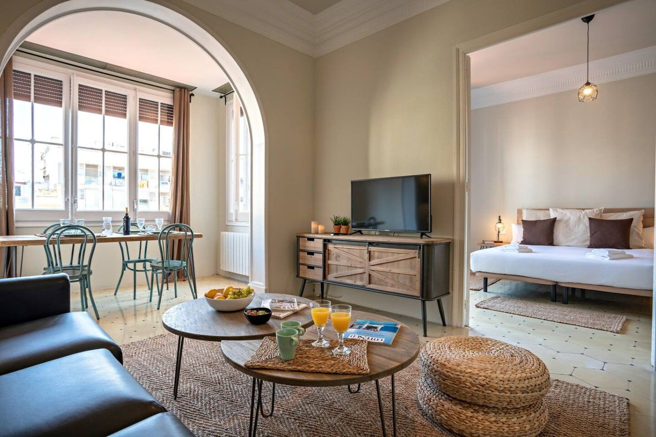 A hotel room with a bed, a TV, a dining area with chairs, a table with breakfast items, and a view through an arched doorway.