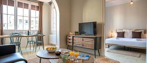 A hotel room with a bed, a TV, a dining area with chairs, a table with breakfast items, and a view through an arched doorway.