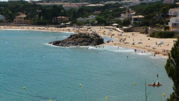 Une scène de plage côtière avec une eau d’un bleu limpide, un promontoire rocheux et une plage de sable où l’on voit des parasols et des gens.