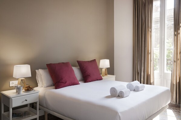 A neatly made bed with white linens, two red pillows, and rolled white towels on a white nightstand with a lamp. - Grace 2 - Habitat Apartments (Barcelona)